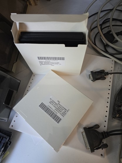 two boxes of 5.25" floppy disks sit on a stack of tractor paper.  one is open showing the disks inside.  to the left are flip top plastic containers with other floppy disks in them.  a serial cable is visible on the right hand side.
