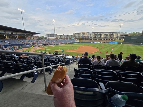 baseball field being prepared for a game.  a hand is holding out a hot dog with a bite taken out.  perspective is from the first base seats about 20 rows back.