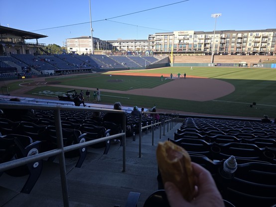 a baseball field from the first base seats.  the field is half in shade.  the grounds crew is making the field ready.  a hot dog is held out in the foreground.