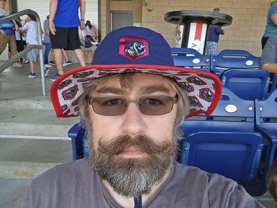 me looking into the camera wearing my new Gwinnett Stripers hat.  it's a wide rimmed floppy hat that is blue in top with the Stripers logo in the middle above the forehead.  it has a white underneath with lots of Stripers logos on it.