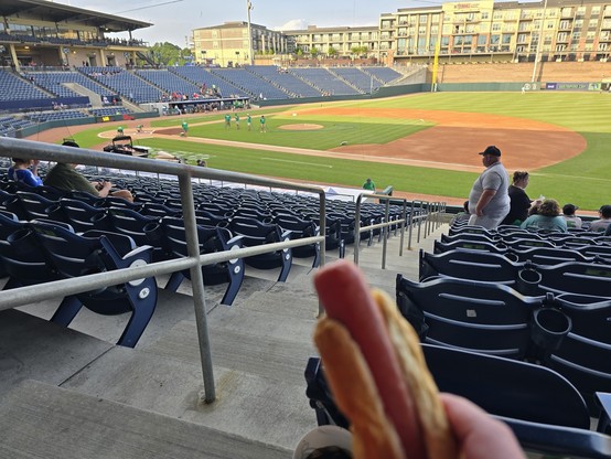 picture of a baseball field being prepared for the game.  perspective is from the first base stands.  in the foreground, a hot dog is visible